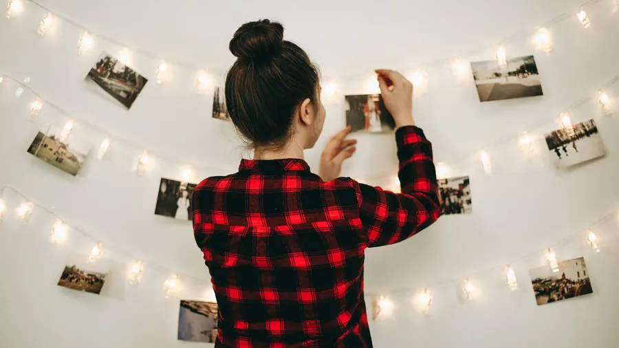 A woman hanging photos on a string light garland using clothespin on a white wall.