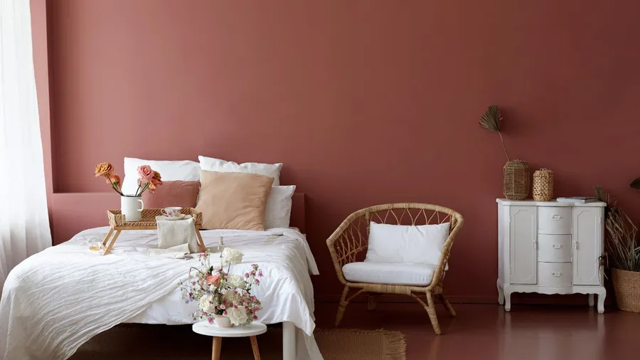 A terracotta-hued bedroom interior with cozy bed, wicker armchair, and a small table with flowers.