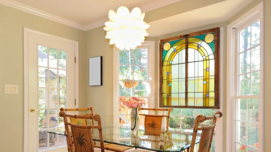 Bright dining area with bamboo-style chairs, a glass table, and a large stained glass window framed by white trim, lit by a sculptural white pendant light and surrounded by tall windows.
