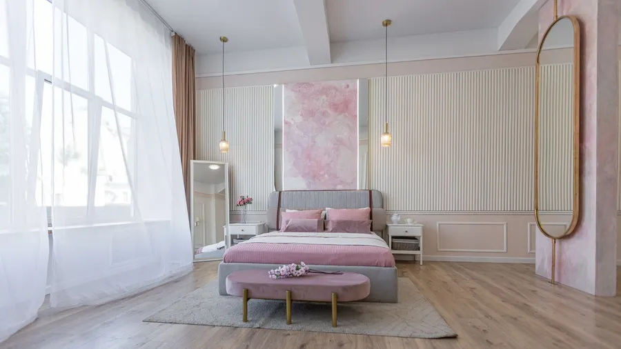 A modern bedroom with a pink and gray color scheme, featuring a bed, nightstands, a large mirror, and decorative lighting.