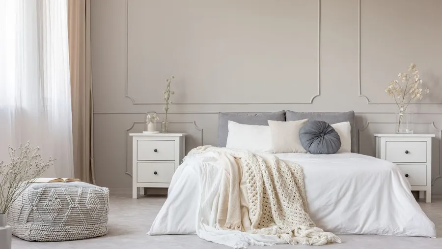 A white bedroom interior with cozy bed with pillows, white throw blanket and chunky knit throw, floor cushion, and white bedside tables.