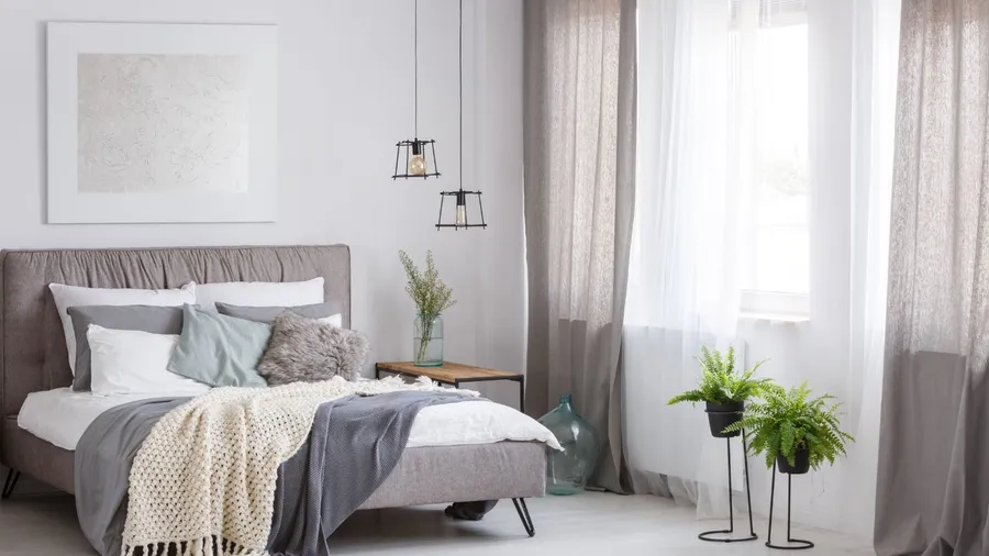 A neutral bedroom interior with a cozy bed, throw blankets, pillows, potted plants, lamps, and layered sheer curtains.