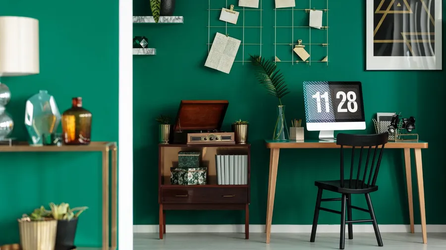 A home office with a green accent wall, featuring a desk with a computer, a record player, plants, and decorative elements.
