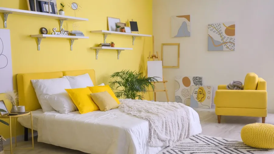 A vibrant bedroom with yellow accent wall with floating shelves, artwork on the wall, yellow armchair, and a cozy bed.