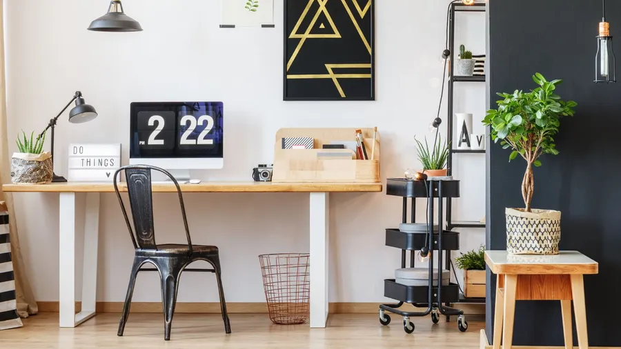 A workspace interior with a wooden table, chair, plant, basket, posters, lamp, and a desk organizer.
