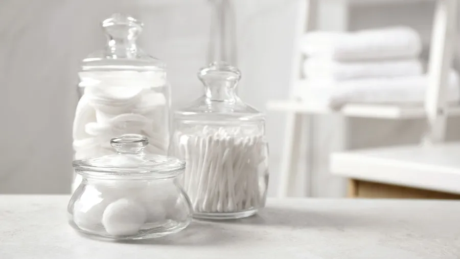 Decorative glass jars with with cotton balls, cotton swabs, and cotton pads on a white countertop in a bathroom.