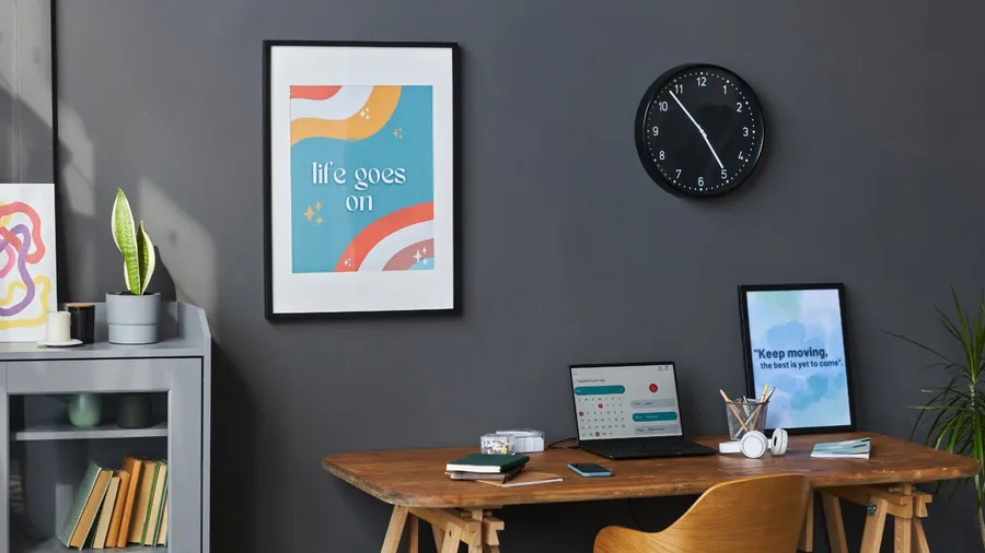 A home office interior with a laptop on wooden table, chair, bookcase, and framed posters on a dark gray wall.