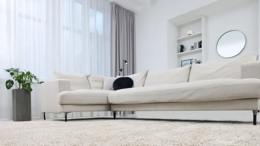 A living room interior with white sectional sofa, rug, houseplant, and floor to ceiling layered curtains.