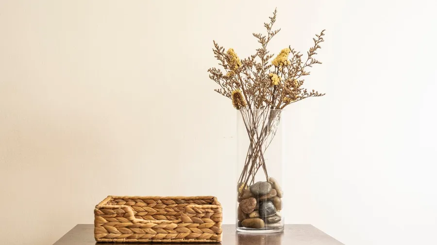 Dried flowers in a glass vase with pebbles and a woven basket on a wooden table.