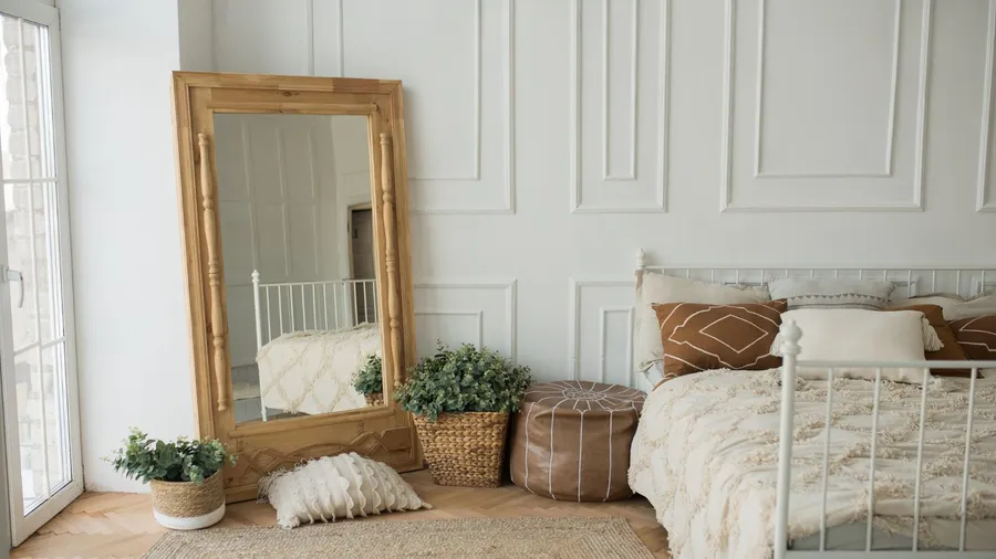 A bedroom interior with cozy bed with earthy brown cushions, brown floor cushion, jute rug, and a full standing mirror with wooden frame near a window.