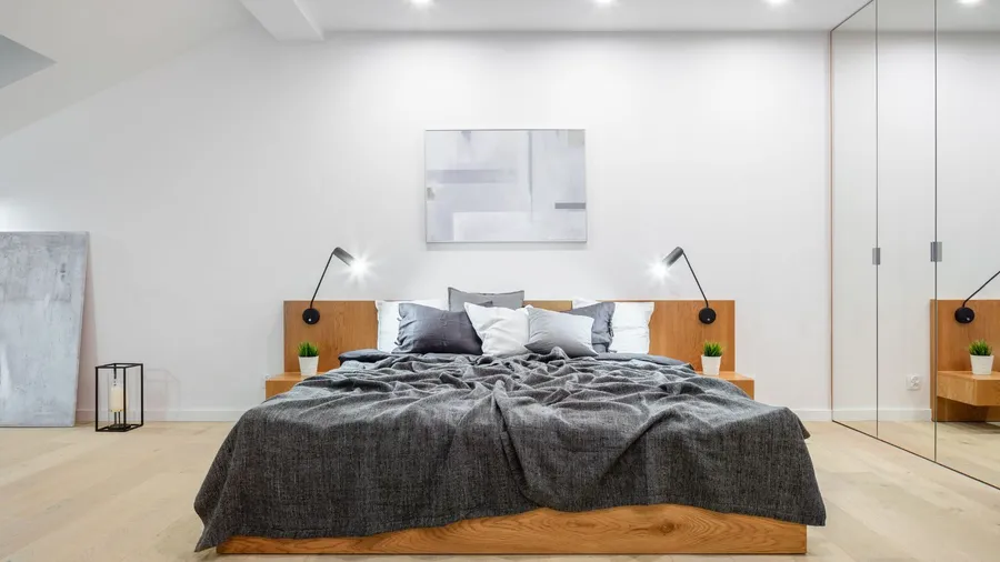 A minimalist bedroom interior with wooden bed with gray bedding, wall art, and mirrored wardrobe.