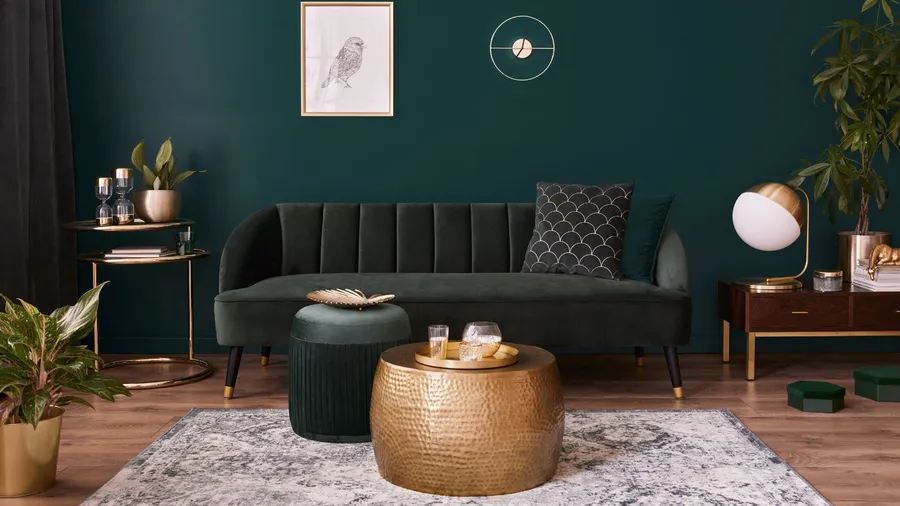 A stylish living room with a dark green velvet sofa, a round gold coffee table, green ottoman, and various decorative items.