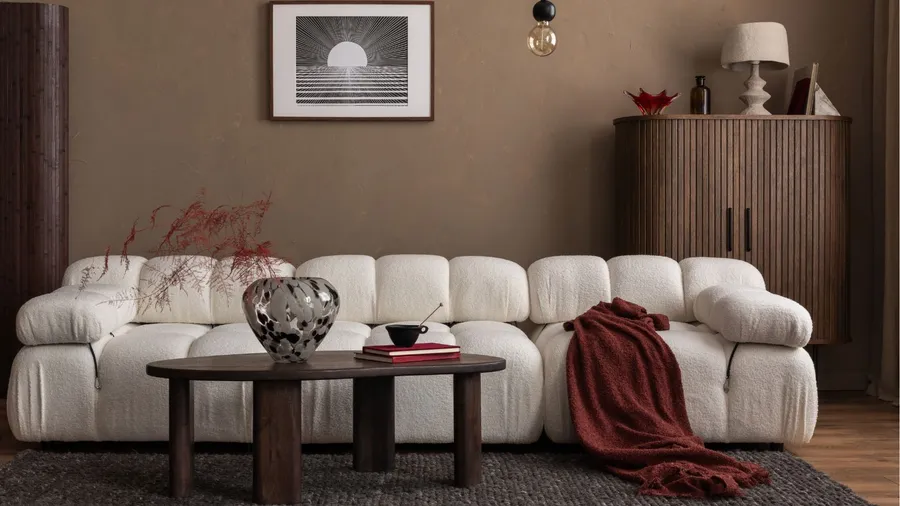 A modern living room features a white modular sofa with a red throw blanket, a dark wood coffee table with a vase and books, and a framed abstract print on a brown wall, with a cabinet and lamp in the background.