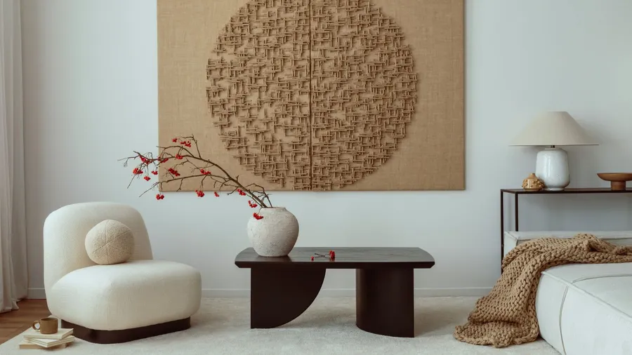 A living room interior with a cozy white sofa, chunky knit throw, wooden coffee table, flower vase, rug, and a large art piece hanging on the wall.