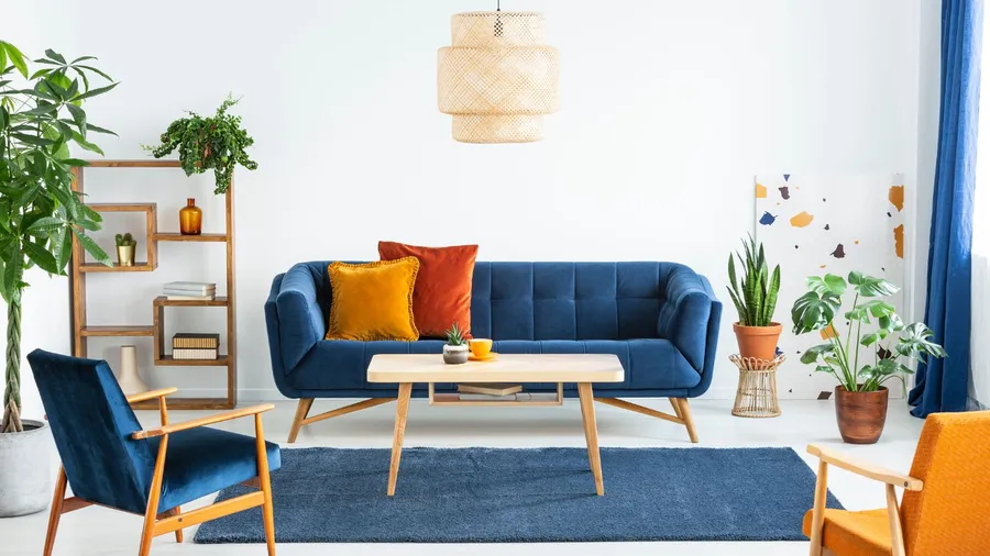 A living room interior with blue sofa with cushions, armchairs, statement ceiling light, plants, coffee table, and a blue rug.