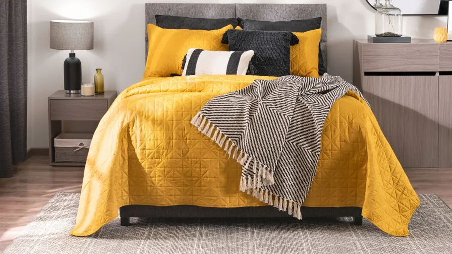 A bedroom interior with a gray bed with yellow and black bedding, quilted yellow comforter, throw blanket, and pillows.
