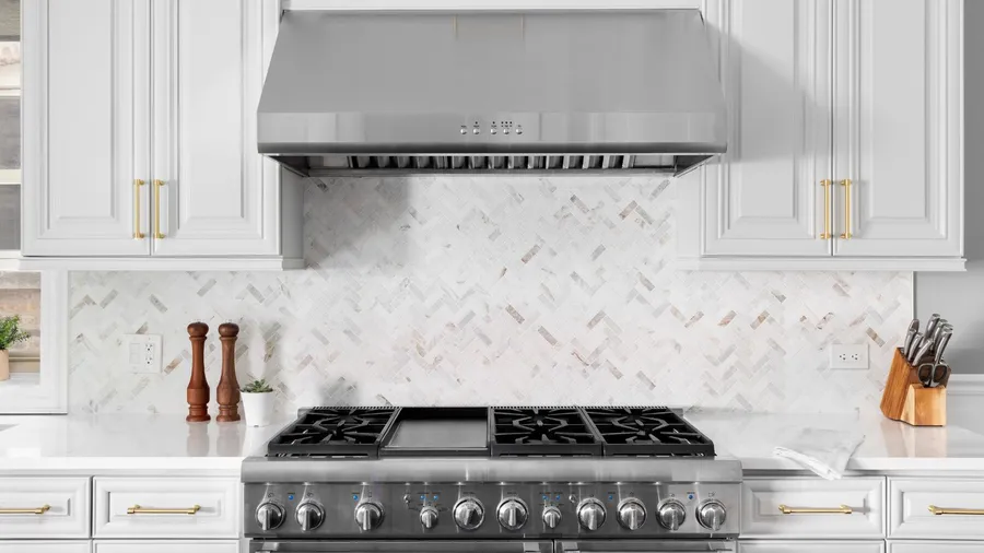 A kitchen interior with white cabinets, stainless steel stove and range hood, granite countertops, and marble herringbone tile backsplash.