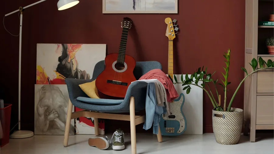 A guitar and clothes on an armchair with sneakers under it in a living room.