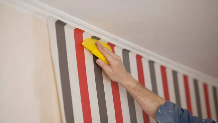 A man gluing colorful vertical stripes wallpaper on a neutral wall.