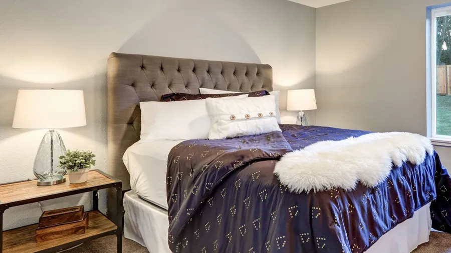 A bedroom interior with a cozy bed with pillows, comforter, and faux fur throw, and a bedside table with lamp and small potted plant.