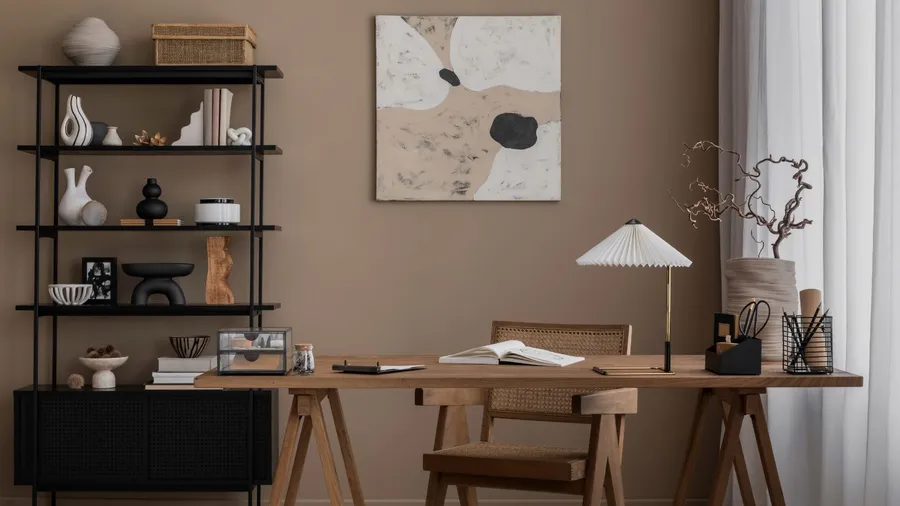 A beige home office interior with wooden table with decorative vase and accessories, chair, black shelf with decor accents, and abstract wall art.