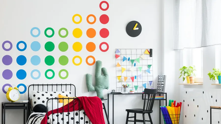 A colorful bedroom interior with a bed with throw blanket and pillows, study table and chair, wall clock, and colorful stenciling on the wall.