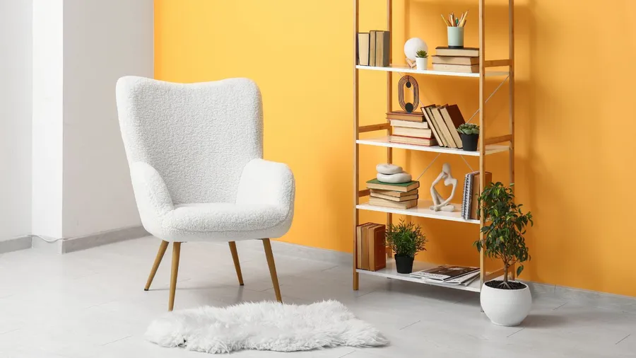 A reading nook with a cozy arm chair, faux fur rug, and a shelf with books and decor accents in front of a yellow accent wall.