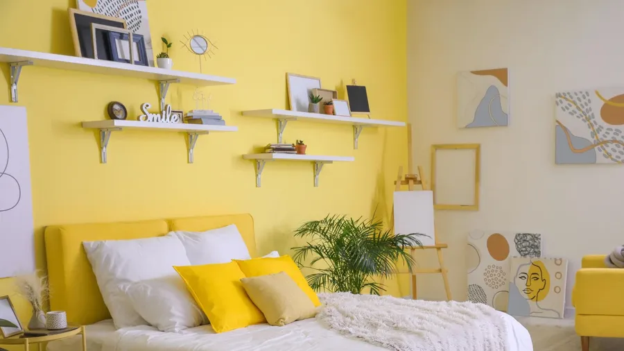 A vibrant bedroom with a bed with pillows and throw blanket, art work, and floating shelves with frames and plants on a yellow accent wall.
