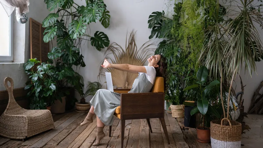 Woman relaxing on a wooden armchair surrounded by various houseplants including monstera, in a cozy, plant-filled room.