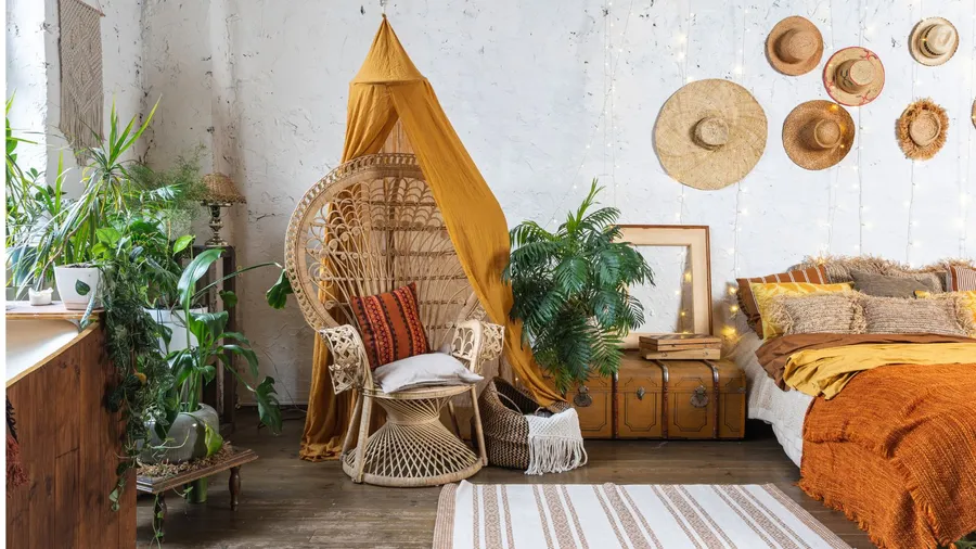 A bedroom interior with cozy bed, wicker chair, potted plants, rug, and vintage chest with books on it.
