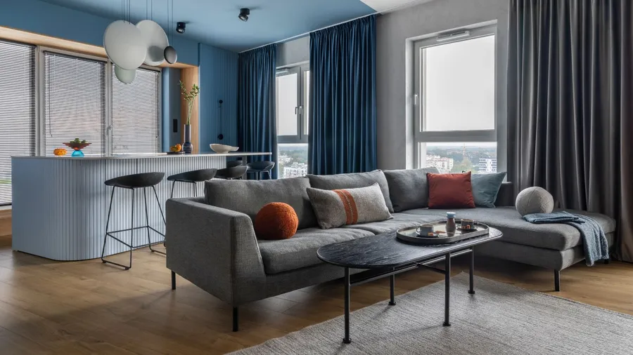 A kitchen and living room interior with a gray sofa, marble kitchen island, black coffee table, curtain, modern sculpture, gray rug, and personal accessories in a luxury apartment.