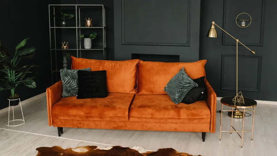 A stylish living room interior with a cozy large orange sofa with cushions, coffee table, floor lamp, houseplants, and a shelf in front of a gray wall.