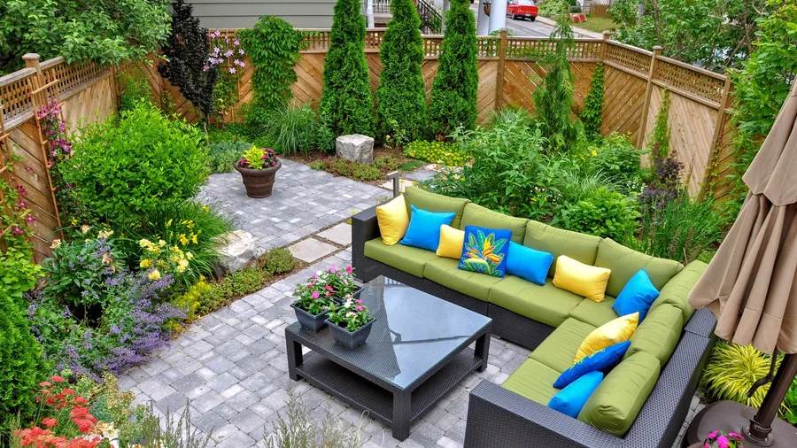 A backyard with a gray sectional sofa with green, blue, and yellow cushions, a table, and colorful plants surrounding it.