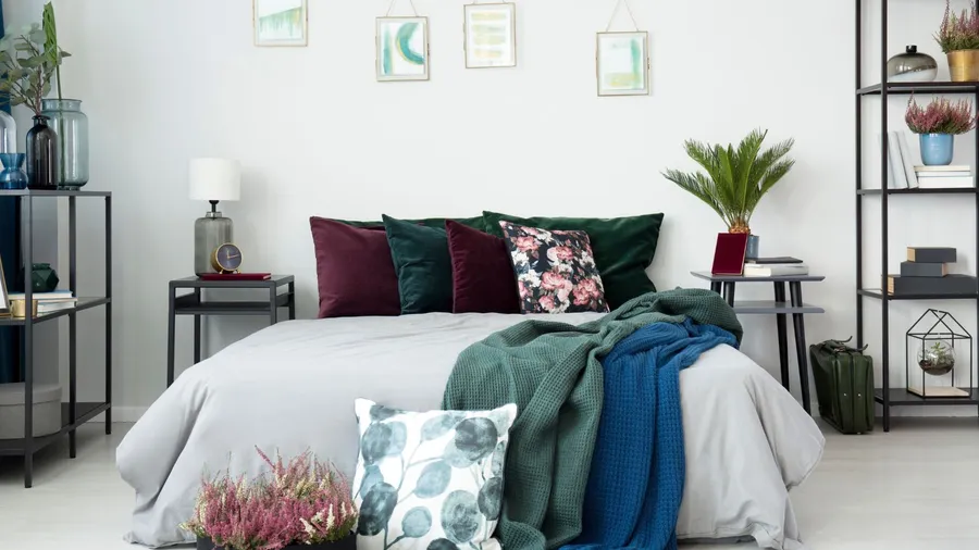 A bedroom with a cozy bed with velvet throw pillows, throw blanket, potted plants, and decor accents on a shelf.