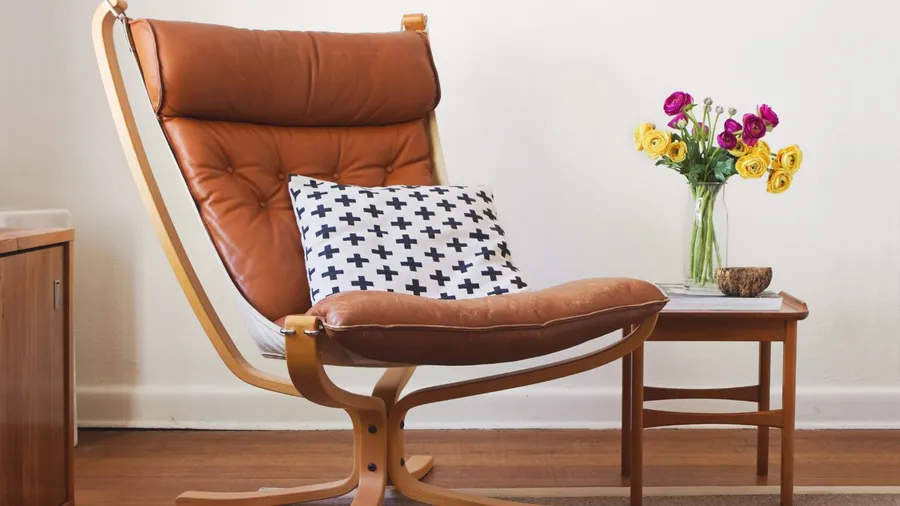 A wooden retro tan chair, wooden coffee table, and a flower vase in front of a white wall.