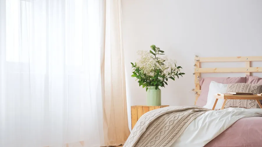 A bedroom interior with wooden bedframe, wooden side table with flowers in a vase, and linen sheer curtains on the window.