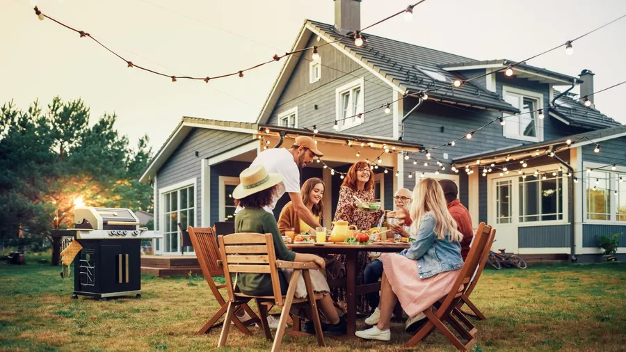 A family enjoying a barbeque party in a garden with illuminated string lights, a grill, and wooden dining setup.