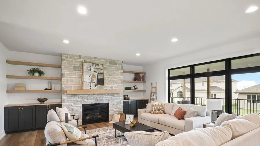 A stylish living room interior with cozy sofas, wooden floating shelves, fire place, and overhead ceiling lights.