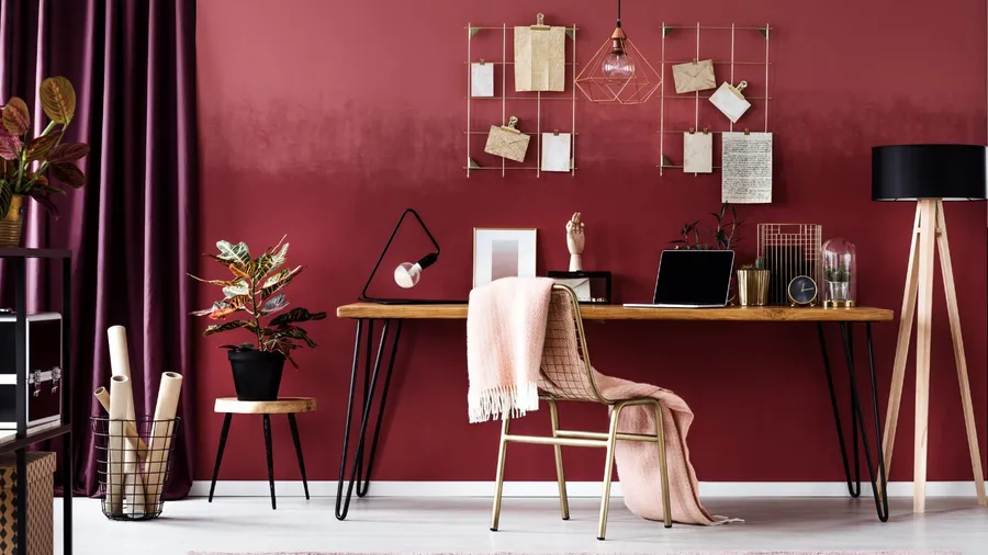 A study zone with a wooden table, chair, throw blanket, lamps, plants, laptop, and mesh grid on a burgundy wall.