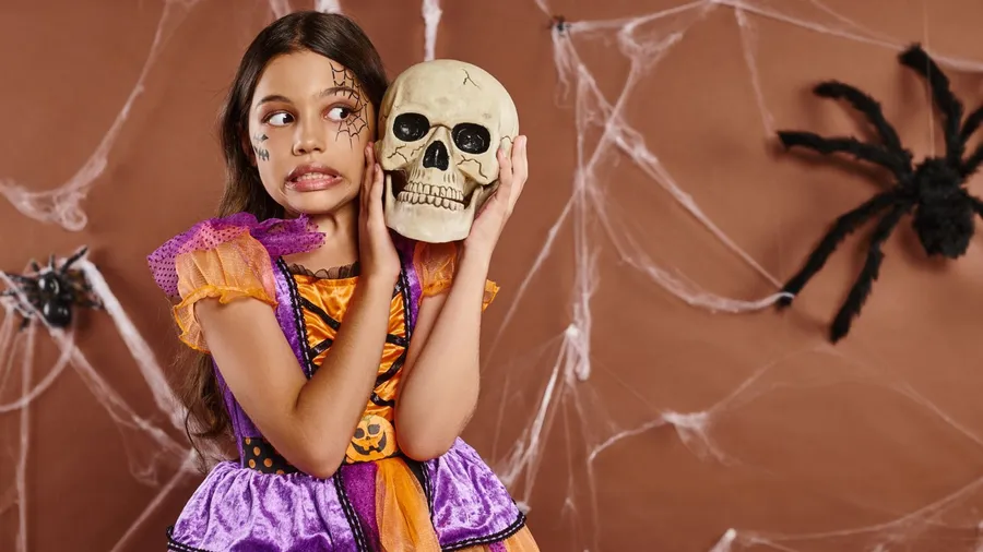 A girl in Halloween attire standing with skull and grimacing on brown background, spooky season.