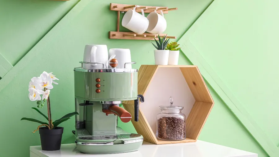 A green coffee machine with stacked cups, surrounded by plants and a jar of coffee beans.