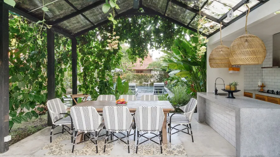 A stylish outdoor dining area with a wooden table, patterned chairs, and a lush green backdrop, featuring a modern open-air kitchen with wicker pendant lights.