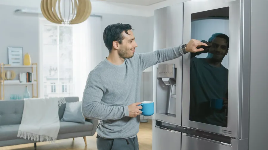 A smiling man is adjusting the settings of a smart refrigerator with built-in ice maker while holding a blue mug.