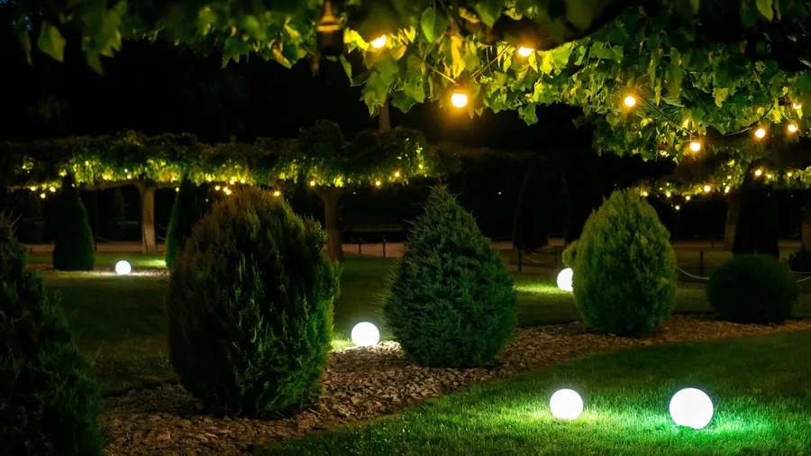 A garden with globe lights on the grass, surrounded by mulch and bushes, with string lights draped over the trees.