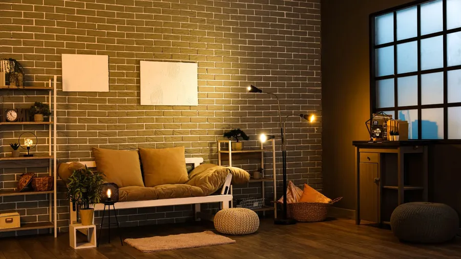 A cozy living room interior featuring a white metal futon with mustard cushions, warm lighting, and a painted brick wall, floor lamp, woven baskets, and a small shelving unit with plants and decor.
