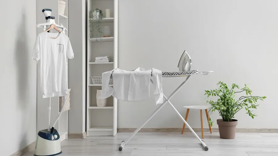 A laundry room interior with a steamer, white shelf, plant, and a foldable ironing board.