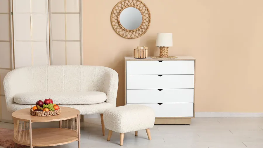 Cozy living room corner featuring a cream-colored armchair and ottoman, a wooden coffee table with fruit, a white dresser, a woven mirror, and a lamp.