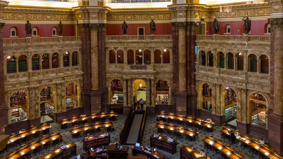 The ornate main reading room of the Library of Congress, featuring rows of desks with lamps, bookshelves lining the walls, and a grand architectural design.