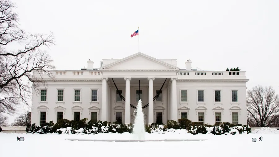 The front view of the White House on a snowy day, with a U.S. flag flying above the roof, a central fountain spraying water, and trees and bushes covered in snow surrounding the building.