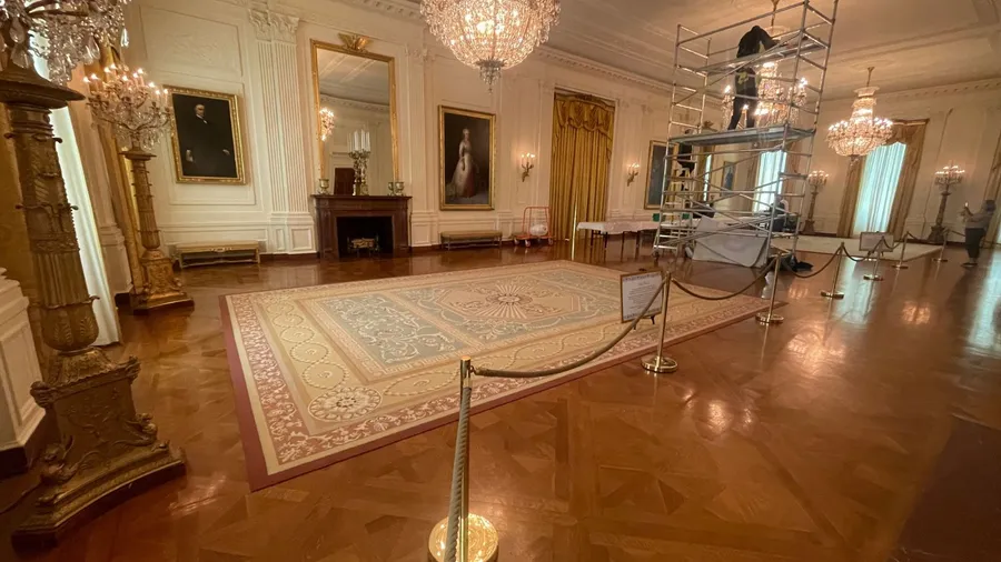 A grand, ornate room, likely in the White House, features chandeliers, large paintings, a fireplace, and a patterned rug, with scaffolding and a worker visible on the right side.